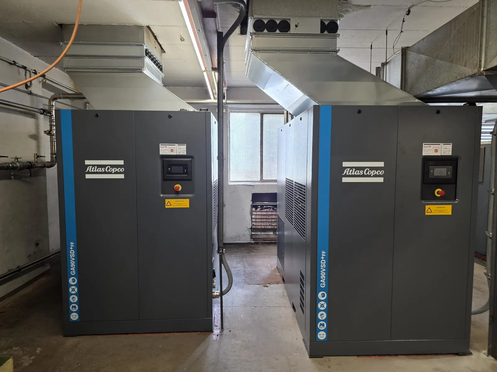 Zwei drehzahlgeregelte Atlas Copco GA 90 VSD+ FF Schraubenkompressoren mit Abluftkanälen in einem Kompressorraum