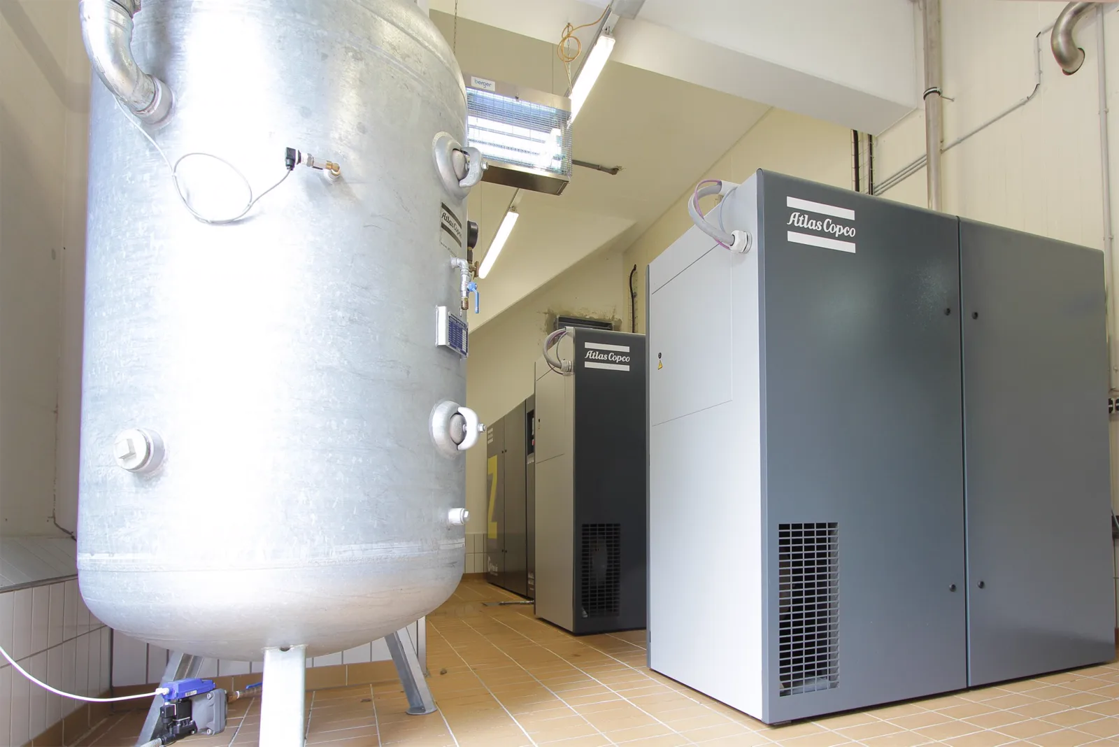Stehender verzinkter Druckluftbehälter mit Manometer und Kondensatablass neben Kompressoren in einem Technikraum